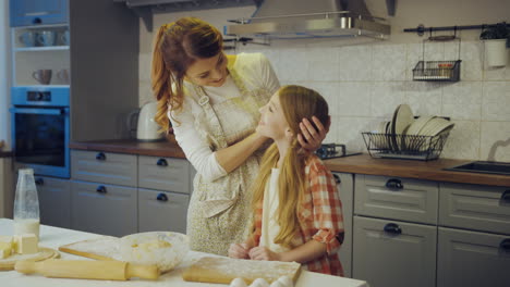 Portrait-shot-of-the-young-mother-and-her-pretty-daughter-caressing-in-front-of-the-camera-and-than-posing-in-the-kitchen-while-making-a-daugh.-Indoors